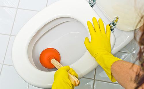 Hands in yellow gloves wash the toilet with a plunger.